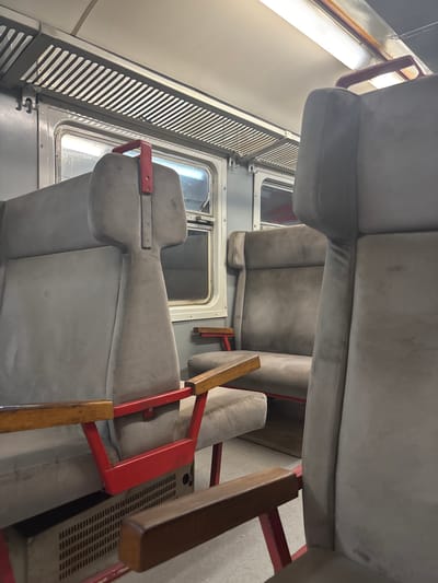 The image shows the interior of a train carriage with empty seats. The seats are upholstered in a light gray fabric and have red metal armrests. There are windows on the side of the carriage, and overhead luggage racks. The scene is dimly lit, suggesting it may be nighttime or the train is underground. This image was taken in Bistrice, Montenegro.