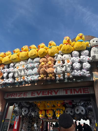 The image shows a close-up, low-angle view of a carnival game stall filled with stuffed animals. The stall is adorned with a large sign that reads "PRIZE EVERY TIME". Rows of yellow duck plush toys are stacked at the top, followed by white and grey bunnies, and then dalmatian dog plush toys. Below the stuffed animals, a dark opening leads to the game area where more plush toys, including monkeys and zebras, are visible. The scene is outdoors with a bright blue sky and scattered clouds in the background. A portion of a person's head is visible in the foreground, indicating someone is viewing or interacting with the stall. The overall impression is one of a lively fairground attraction. The stall appears to be located in a fairground or amusement park, with the mention of "Borough of Runnymede, United Kingdom" suggesting a specific geographical location for this scene.