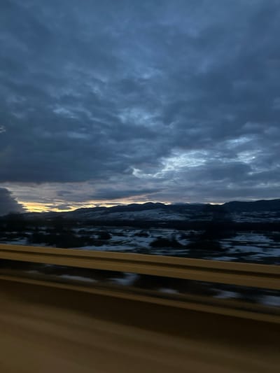 An eerie dusk scene unfolds from what appears to be a window. A body of water reflects the dark, clouded sky with a strip of orange sunset glow on the horizon. The land appears as a dark silhouette. Window slats are visible at the right. The dominant mood is one of dramatic, impending weather.