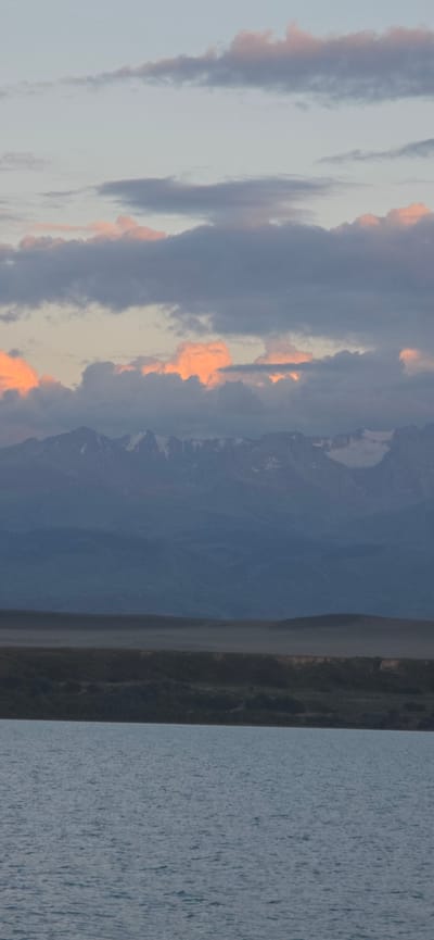 The image depicts a serene landscape with a body of water in the foreground, reflecting the soft hues of the sky. In the mid-ground, a gently sloping hill is visible, leading towards a majestic mountain range in the distance. The mountains are capped with snow, adding a dramatic contrast to the earthy tones of the terrain. The sky is adorned with a scattering of clouds, some tinged with the warm glow of sunset or sunrise, while others are a soft, muted gray. The overall scene evokes a sense of tranquility and natural beauty. The location is identified as Pristan'-Przheval'sk, Kyrgyzstan, suggesting a setting within this region. No people or specific man-made structures are visible in the image, focusing entirely on the natural elements.