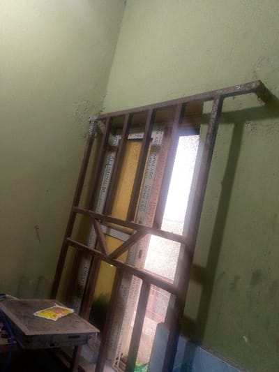A metal frame leans against a wall with a faded, yellow tint. In the background, window panes bear repetitive text including "OSY PAPA" and "ALUMINIUM." A small, flat object rests on a wooden platform in the foreground. The wall exhibits uneven texture and coloration, transitioning from yellow to a greenish hue near the floor. The lighting is uneven, casting shadows and emphasizing the imperfections in the wall's finish.