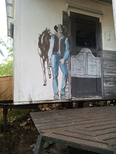 A building features a mural of a cowboy standing next to a horse. The cowboy wears a hat, vest, and blue jeans. To the right of the mural, a door with a small window and saloon-style lower half is visible. The building appears to be raised off the ground, supported by a metal frame. A wooden deck is situated in front of the building, with visible gaps between the planks. Green vegetation is present in the surrounding area.