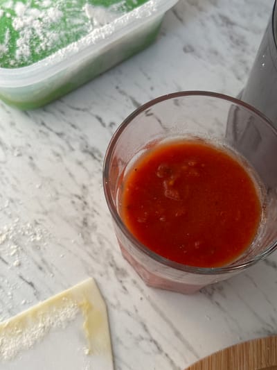 A close-up, high-angle shot shows a glass of red sauce and a plastic container with a green substance dusted with white powder, both on a marble-patterned countertop. A small piece of dough, dusted with flour, is visible in the lower left corner. A blurred dark object, possibly a blender, is in the background on the right. The scene appears to be in a kitchen. The image does not contain information to locate it in Moortown, Ireland.