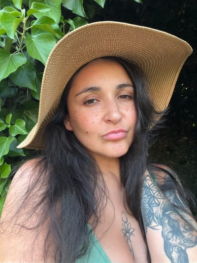 The image shows a close-up, waist-up shot of a woman wearing a wide-brimmed straw hat and a green top. She has long, dark hair and visible tattoos on her left arm and chest. Her face is tilted slightly, and her lips are pursed. The background consists of lush green foliage, likely ivy, and dark shadows, suggesting an outdoor setting. The location is specified as Ароматненское сельское поселение, Ukraine.