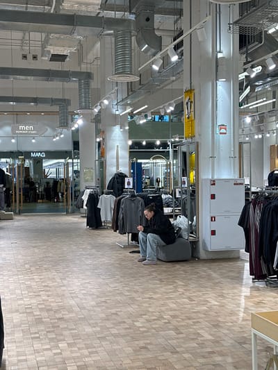 A woman in a black jacket and jeans browses clothes in a store. She leans over a rack of garments. Clothing hangs on racks around her. The store has exposed ductwork and lighting fixtures. A "men" sign is visible. The floor has a wood grain pattern. A large white box sits behind the woman.
