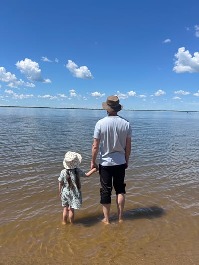 In the city of Telmana, Russia, a man and a young girl stand in shallow water, holding hands and looking out at a wide body of water under a blue sky with scattered white clouds. The man is wearing a light-colored t-shirt, black shorts, and a brown hat. The girl is wearing a light-colored dress with a pattern and a matching hat. The water is murky brown near the shore, with ripples reflecting the sunlight. In the distance, a faint red buoy is visible on the water. The shoreline is sandy and appears to be a natural environment.