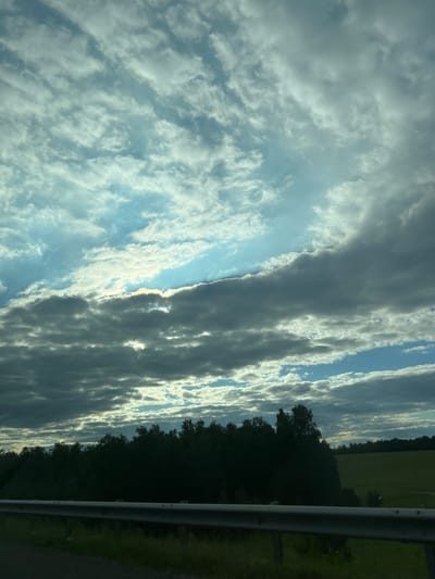 The image captures a cloudy sky over a rural landscape, with a silver guardrail in the foreground. The sky is filled with a mix of dark, heavy clouds and lighter, wispy formations, with patches of blue sky visible. Sunlight streams through some of the cloud gaps, creating a dramatic effect. Below the sky, a dense line of dark green trees forms a silhouetted backdrop. To the right of the trees, a green, open field is visible, suggesting a rural or agricultural setting. The guardrail runs horizontally across the lower portion of the image, indicating a road or highway. The ground immediately behind the guardrail appears to be covered in grass and some darker earth. The lighting suggests it is either early morning or late afternoon. The image does not contain any people or visible signs of recent activity. The location is identified as Sukhaia Balka, Russia, which is a rural area.