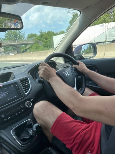 The image depicts the interior of a Honda car, focusing on the driver's area. A person is shown driving, with their hands on the steering wheel. The gear stick indicates a manual transmission vehicle.
The dashboard is visible, including the instrument panel, the radio, and the air conditioning controls.
The location is inferred to be Milton Keynes, United Kingdom based on the "Call 0800 521 728" printed on the windshield which is a United Kingdom emergency line.
Through the windshield, a view of the outside environment can be seen, including buildings and vegetation.