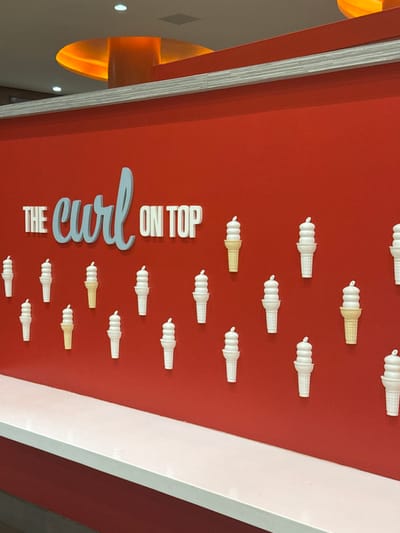 An interior wall of an establishment in Boca del Río, Mexico, is depicted. The wall is painted a solid red. Mounted on the upper central portion of the wall are three-dimensional letters spelling "THE curl ON TOP," with "THE" and "ON TOP" in white block letters and "curl" in a light blue script font. Below the text, approximately two dozen decorative ice cream cones, depicting soft-serve ice cream, are affixed to the wall in a staggered pattern. Most cones are white with white soft-serve, while a few are light yellow/tan with white soft-serve. A white horizontal counter or ledge extends across the lower portion of the image, positioned in front of the red wall. Above the red wall, a silver-gray horizontal trim strip separates it from a white ceiling. Recessed lighting fixtures are visible in the ceiling, along with a prominent circular architectural feature that is illuminated with an orange glow in the upper left. No individuals or active interactions are present in the scene.
