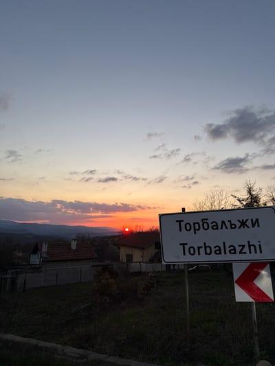 The image depicts a sunset over the city of Torbalazhite in Bulgaria. In the foreground, a white road sign with the city's name in Cyrillic and Latin script is visible. A red and white chevon sign is attached above the city sign. The sky transitions from gray to orange and red as the sun sets behind distant hills. The outlines of houses and buildings are visible below the horizon. Bare trees are silhouetted against the sky. The foreground contains vegetation and parts of a wall.