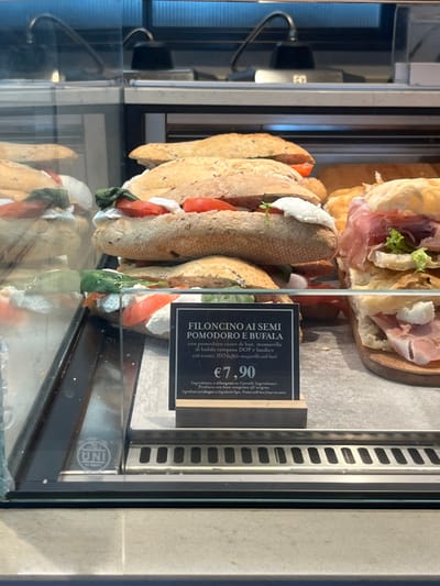The image shows a well-lit food display case, likely in a deli, cafe, or bakery. The perspective is from slightly above, looking into the glass counter where various sandwiches are presented for sale. The overall scene suggests a commercial food establishment.
On the upper shelf of the display, two sandwiches filled with thinly sliced prosciutto and green lettuce are visible, resting on a wooden board. Below these, and more prominently in the foreground, are several long, baguette-style sandwiches, cut open to reveal their fillings. These sandwiches feature thick slices of red tomato, generous portions of white mozzarella cheese, and fresh green basil leaves, encased in seeded bread.
A black price and description sign stands in front of the baguette sandwiches. The text, predominantly in Italian with some English translation, reads:
"FILONCINO AI SEMI POMODORO E BUFALA" (Small seeded baguette with tomato and buffalo mozzarella)
"con pomodoro cuore di bue, mozzarella di bufala campana DOP e basilico" (with oxheart tomato, PDO buffalo mozzarella and basil)
"with tomato, PDO buffalo mozzarella and basil" (English translation)
The price displayed is "€7,90". Additional text states: "Ingredienti e allergeni su Cartelli Ingredienti." (Ingredients and allergens on Ingredient Cards.) and "Prodotto con base congelata all'origine." (Product made from frozen base at origin.)
The interior of the display case is bright, suggesting either ample natural light or strong artificial lighting. Reflections are visible on the glass, and a faint "UNI" logo can be seen on the metallic frame of the display case in the lower right corner. The specific food items and Italian language cues indicate a location in Italy, consistent with Dorno.