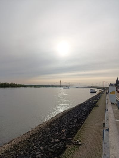 The image captures a scene in Emmerich on the Rhine, Germany. A wide river dominates the left side, with sunlight glinting on its surface. A bank of dark, jagged rocks lines the river's edge. Adjacent to the rocks is a paved walkway, its surface marked with patterns. A metal railing runs along the walkway's edge. In the distance, a red suspension bridge spans the river, with several boats visible on the water. On the right side, a church tower rises above buildings near the walkway. The sky is overcast, with the sun obscured by a thin layer of clouds.