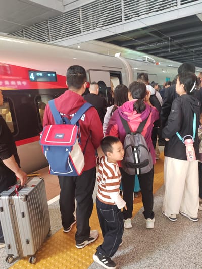 The image depicts a busy scene on a high-speed train platform in Yuhuatai District, China. A crowd of people, including adults and a child, are gathered on the yellow tactile paving, seemingly waiting to board a modern red and white train.
The train, a sleek high-speed model, is docked at the platform. A digital display on its side clearly shows "4 G740 16-01," indicating train number G740, possibly car 16 and door 01. The platform is well-lit, suggesting either daytime or bright indoor lighting conditions. Above the train and passengers, the station's overhead structure with horizontal grates is visible.
In the foreground, a person wearing a red hoodie and a prominent blue and red backpack with "PUXIN" written on its flap stands with their back to the viewer. To their left, a silver, hard-shell rolling suitcase is present. A small child, dressed in a brown and white striped long-sleeve shirt and dark pants, stands near the red-hoodied individual, looking towards the right side of the frame. Further to the right, another person in a purple and pink jacket wears a gray backpack, while an individual in a black jacket and white wide-leg pants carries a pink water bottle with a light blue strap, featuring a cartoon bunny and the text "ANDY DINO." The overall impression is one of a bustling travel hub as passengers prepare to embark on their journey.