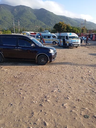 This image captures a bustling, unpaved matatu (commuter van) stage in Namanga, Kenya, set against a backdrop of lush, green mountains under a partly cloudy sky. The scene appears to be taking place during the late morning or early afternoon, characterized by bright, natural light and moderate shadows.
In the foreground on the left, a dark-colored SUV or minivan, possibly a Toyota Sienta, with tinted windows and custom wheels, is parked facing right. The immediate foreground consists of rough, sandy-brown dirt and gravel, showing tire tracks and small stones.
The mid-ground is dominated by several white matatus, which are common public transport vehicles in Kenya. These vans feature blue and orange branding. One prominent matatu in the center-right clearly displays "NAEKANA SACCO" on its front and side. Another matatu behind the dark SUV on the left has "IMANI EXPRESS" and "MBALAA 8000" written on its side. A sign further back, above the matatus, partially shows "NAIROBI," suggesting a destination or transport company name like "Nairobi Executive" (as seen on some matatu branding).
Several individuals are visible around the matatus, suggesting activity related to public transportation. One person in a light-colored uniform stands near the central "NAEKANA SACCO" matatu, while others mill about, possibly waiting for rides or attending to the vehicles.
In the background, tall, green-covered mountains create a dramatic natural setting. Utility poles and communication towers are also visible on the left, blending into the landscape. On the far right, small, rustic structures or kiosks are partially visible. The overall scene depicts a functional and active transportation hub in a rural or semi-urban environment.