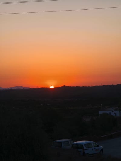 The image captures a sunset over a rural landscape, possibly in the vicinity of Riba-roja de Túria, Spain. The sky is a gradient of orange and pale yellow, with the sun a bright, partially obscured disc just above the horizon line. In the mid-ground, a dark, silhouetted hill or ridge spans the width of the frame, dotted with several tall, thin structures that appear to be power line pylons. The foreground is dominated by dark foliage and two parked vehicles. One vehicle, a light-colored van, is positioned on the right side of the frame, facing left, with a logo visible on its side. Another light-colored vehicle, possibly a van or SUV, is partially visible to the left of the first vehicle, also parked. The overall scene suggests a quiet, twilight setting in a natural environment.