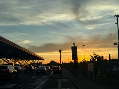 The image depicts a sunset scene at what appears to be an airport drop-off and pick-up zone in Uttlesford, United Kingdom. A line of cars is queued on a wet road in front of a modern building with a large, slanted roof structure, likely an airport terminal. The sky is filled with dramatic clouds illuminated by the setting sun, casting a warm orange and yellow glow. Streetlights and a tall pole with multiple lamps are visible, along with a sign labeled "Zone B Drop Off & Pick Up". A person is seen walking on a paved walkway beside the road, pulling a suitcase, silhouetted against the bright sky. The environment suggests a travel hub during a busy period at dusk.
