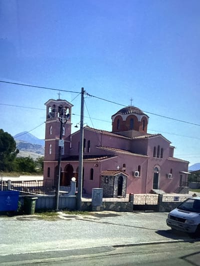 The image shows an exterior view of the Church of Agios Georgios in the village of Myrodato, Prosotsani Municipal Unit, Greece. The church is pink with a bell tower on the left and a dome on the right. It has arched windows and entrances. There is a small stone shrine in front of the church. In the foreground, there is a road, a fence, and some garbage bins. A light-colored car is parked on the right side of the road. In the background, there are mountains and trees. The sky is clear and blue.