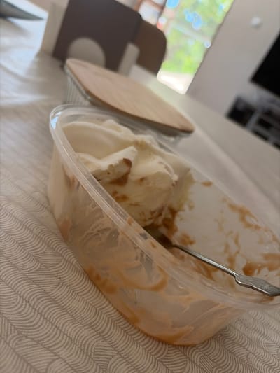 The image shows a partially eaten container of ice cream with a spoon. The ice cream appears to be vanilla with a caramel swirl. The container is clear plastic, and streaks of ice cream and caramel are visible on the inside walls. A silver spoon with a decorative handle is resting in the ice cream. To the left of the ice cream container is a wooden lid or serving board. The scene is set on a white textured surface, possibly a tablecloth or bedsheet, with a wavy pattern. In the background, out of focus, are hints of a room, including what looks like a window with sunlight streaming through, showing blurred greenery and possibly blue sky, suggesting daytime. There is also a dark, out-of-focus object that might be a piece of furniture or electronic device. The overall impression is a casual, indoor setting. No text is visible in the image. The context provided indicates the location is Kukorevo, Bulgaria, but this is not visually discernible from the image itself.