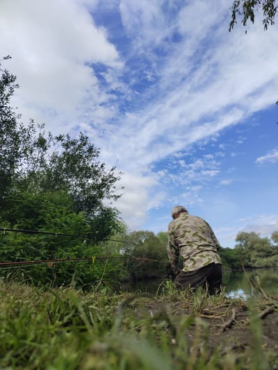 A lone fisherman wearing a camouflage shirt and dark pants is positioned by the edge of a body of water. Several fishing rods are set up in the foreground, extending towards the water from behind a dense green bush. The water reflects the bright blue sky, which is filled with scattered white clouds. Lush vegetation, including trees and tall grass, surrounds the water and the fisherman. The perspective is low, with the grass in the immediate foreground appearing out of focus. The location is identified as Krushare, Bulgaria.