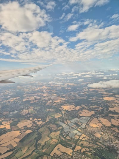 An aerial view captured from an airplane window shows a vast landscape of agricultural fields and scattered buildings under a partly cloudy blue sky. The airplane's wing is visible in the upper left quadrant of the image, extending into the frame. The land below is a patchwork of tan and green, indicating different crops and vegetation, with a river or stream meandering through the terrain. A cluster of buildings, resembling a town or village, is visible in the lower half of the image, surrounded by fields. The sky is a mix of bright blue and white cumulus clouds, casting shadows on the land below. There are no people or specific objects that would allow for the identification of the city of South River, United States of America.