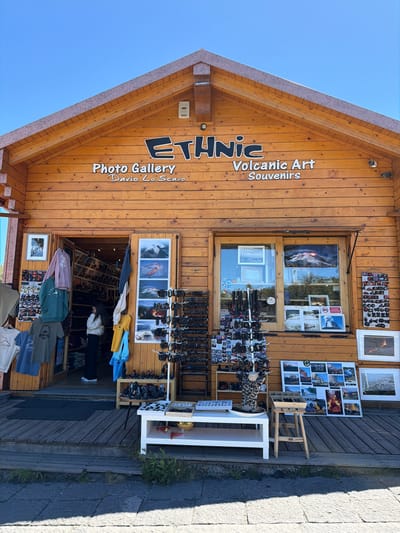 The image shows the exterior of a wooden shop with a prominent sign that reads "ETHNIC Photo Gallery Dario Lo Scavo Volcanic Art Souvenirs". The shop is located in Linguaglossa, Italy, a town known for its proximity to Mount Etna.

The shop's facade is made of horizontal wooden planks. Above the entrance, the "ETHNIC" logo is in large, raised letters, with "Photo Gallery Dario Lo Scavo" and "Volcanic Art Souvenirs" written below it in smaller text. There are two windows and an open doorway leading into the shop.

On the left side of the entrance, several t-shirts and hoodies are displayed on hangers. Inside the open doorway, a young woman with dark hair, wearing a white sweater and dark pants, stands near shelves stocked with photographs.

To the right of the doorway, a window displays various items. Below the window and to the right of the entrance, a variety of merchandise is displayed on shelves and tables. This includes racks of bracelets, sunglasses, framed photographs, and collage prints of volcanic landscapes. A white table in front of the shop holds a display of items, possibly souvenirs or jewelry.

The shop is situated on a wooden deck with steps leading down to a paved area. Green plants grow along the edge of the paved area. The sky above is clear and blue. The overall scene suggests a tourist-oriented shop selling art and souvenirs related to volcanic activity, likely Mount Etna given the location.