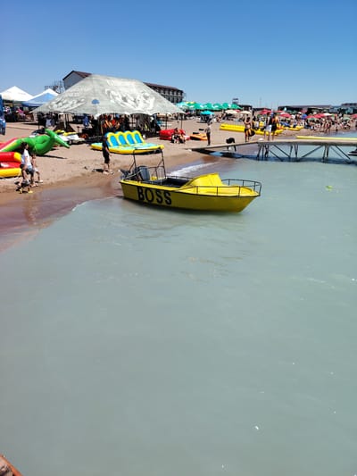 The image shows a crowded beach scene at Lake Kapchagay, located near the village of Toranghylyk, Kazakhstan. The day is clear and sunny. In the foreground, a yellow boat with "BOSS" written on its side is partially submerged in the pale blue water, near the sandy shore. Several inflatable water toys, including a green crocodile and yellow and blue loungers, are visible on the beach. People are seen on the sand, some walking and others relaxing under umbrellas. A wooden pier extends into the water, with individuals standing on it. In the background, buildings and more umbrellas dot the shoreline, indicating a popular recreational area.