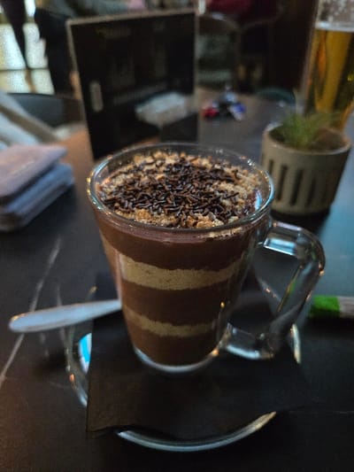 A clear glass mug containing a layered dessert is centrally positioned on a dark, reflective table, possibly patterned with faint white veining, in Negorci, North Macedonia. The dessert exhibits alternating layers of dark brown and light brown substance, topped with a layer of light-colored crumbs and sprinkled with dark brown chocolate sprinkles. The mug has a clear handle and rests on a small, dark, round saucer, with a folded black napkin placed beneath the mug, partially covering the saucer. To the immediate left of the mug, a silver spoon lies on the table. In the blurred background to the left, a stack of light-colored, folded cloths is visible. Directly behind the mug, a dark, rectangular menu or stand is discernible. To the right, a small potted plant in a light brown, ribbed pot is present, with a tall glass containing an amber liquid, consistent with beer, positioned behind it. Farther in the background, indistinct, out-of-focus figures indicate the presence of other individuals within an indoor public establishment. No specific actions or direct interactions are discernible in the static scene.