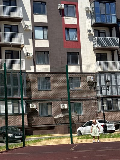 In the city of Vyazniki, Russia, a young girl in a light green dress stands on a red-colored court, likely part of a sports complex, behind a chain-link fence. She is looking towards the camera. To her right, a white car is parked on a grassy area. Behind the fence and the grassy area, a multi-story brick building with numerous windows is visible. The building has air conditioning units mounted on its exterior. In the foreground, the red court is marked with white lines. The scene appears to be during daylight, with shadows cast by the building and the fence. A black car is parked on the street in front of the brick building.