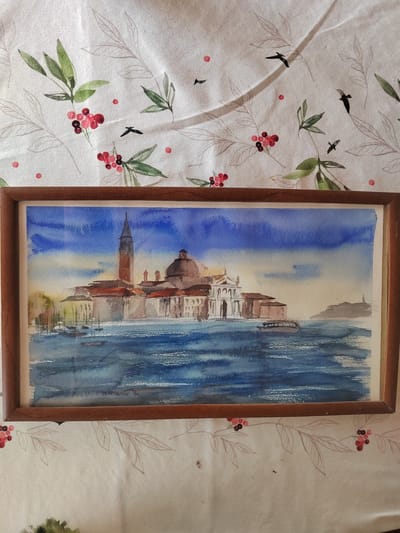 The image shows a framed watercolor painting of a cityscape with water. The painting depicts buildings with a tall bell tower and a dome, situated along a body of water. Boats are visible on the water. The sky is painted in shades of blue and yellow. The painting is framed in a wooden frame. The painting is placed on a white tablecloth with a pattern of green leaves and red berries, and silhouetted birds. The question mentions the city of Бярэзінскі сельскі Савет, Belarus; however, the painting itself does not depict this location.
