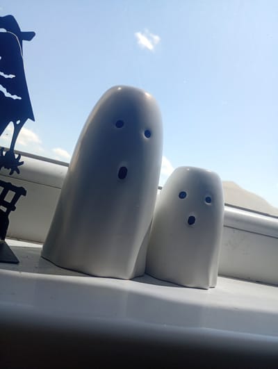 Two white ceramic ghost figures sit on a white windowsill. The larger ghost on the left has two black dots for eyes and an oval black mouth. The smaller ghost on the right has two black dots for eyes and a black oval mouth. A black silhouette of a witch on a broomstick is visible to the left of the ghosts. The background shows a bright blue sky with a few white clouds. The scene is indoors, with the windowsill appearing to overlook an outdoor environment, possibly a residential area in Aidemir, Bulgaria.