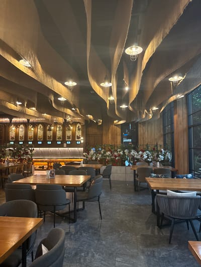 This is a vertical, full shot of a restaurant interior. The ceiling is a striking architectural feature, with large, undulating fabric panels in a neutral tone, accented by hanging industrial-style pendant lights. The walls are paneled with wood, adding warmth to the space.
The restaurant is furnished with modern, dark gray upholstered chairs and wooden tables. In the background, a bar area is visible, lined with bottles and illuminated by warm lighting. Decorative elements include arrangements of red and white flowers on a railing and framed artwork or menus on the wall. The flooring appears to be large, dark gray tiles.
The scene suggests a calm and inviting atmosphere, likely in the evening or indoors, as the lighting is prominent. The overall aesthetic is contemporary with a touch of industrial chic. There are no people visible in the frame. The location cues of Ban Na Chom Thian, Thailand, are not directly evident in the visual elements of the image itself. Some small framed prints are visible on a table in the foreground.