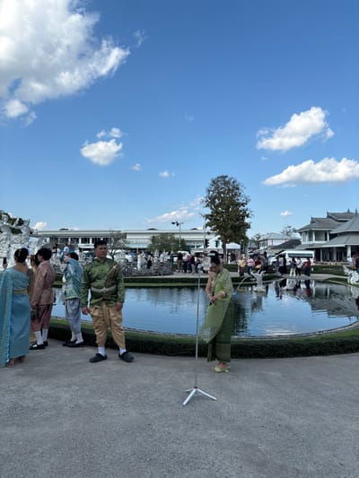 The image depicts an outdoor scene at Wat Rong Khun, located in Ban Rong Khun, Chiang Rai province, Thailand, under a clear blue sky with scattered white cumulus clouds. In the foreground, a paved grey surface occupies the lower third of the frame.
Central to the midground is a dark-surfaced, irregularly shaped pond with manicured green hedges bordering its edge. Several ornate, white, sculptural elements, typical of the temple's architecture, are visible within and around the pond, with some functioning as fountains.
Approximately eight individuals in traditional Thai attire are visible in the midground and left midground. A man wearing a green and gold patterned traditional top with beige trousers and black slip-on shoes stands left of center, looking towards the right. To his left, several individuals, including a woman in light blue and gold attire and another in light pink and gold, are standing near the pond's edge. To the right of the central man, a woman in a light green patterned traditional dress stands next to a white tripod with an extended pole, appearing to interact with or adjust a device attached to it. She wears light green slip-on shoes.
In the background, a large building with a long, white facade and numerous windows is visible, stretching across the width of the frame. Further right, traditional Thai-style buildings with dark roofs are apparent. Numerous tourists in casual clothing are scattered throughout the background, congregating near buildings and around the pond. A prominent, dark-leafed tree stands centrally in the mid-to-background, obscuring part of the distant architecture. Streetlights and other utility poles are also visible in the background.