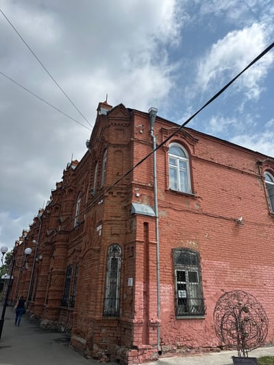 In Nogornyi, Russia, an old red brick building with ornate architectural details lines a street. The building features arched windows, some with decorative metal grates, and elaborate brickwork along its facade. A person is walking away from the camera down the sidewalk next to the building. The sky above is partly cloudy with a mix of blue sky and gray clouds. Power lines are visible stretched across the sky. A circular metal sculpture with a fruit-like design sits in front of the building. Security cameras are mounted on the building's exterior.