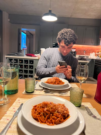 A man with curly dark hair wearing a grey cable knit sweater is seated at a wooden table, holding a smartphone in his hands and looking at it. On the table in front of him is a plate of spaghetti with what appears to be meatballs and sauce. There is a wine glass and a green textured glass to his right. Another place setting with a similar plate of spaghetti, a fork, knife, and another green textured glass is visible to the right of his plate. In the background is a kitchen area with grey cabinetry and an orange tiled backsplash. There is a wine rack behind the man containing a number of bottles. A hanging light fixture is visible above the man. There is a hint of a window or opening in the wall behind the wine rack. There is no information about the location of the city Gondarém, Portugal, visible in the image.