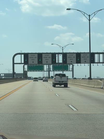 An eye-level, medium shot captures a view of traffic moving on a highway in Oxon Hill, Maryland. Overhead signs display "STOP HERE ON RED" in white lettering against black backgrounds, with downward-pointing arrows. Green traffic lights are illuminated below these signs. A white pickup truck is the closest vehicle, followed by a white SUV and a dark-colored SUV. The road is paved, with a yellow line marking the left lane and white dashed lines separating lanes. The sky is a clear blue with scattered white clouds, and streetlights are visible on both sides of the highway.