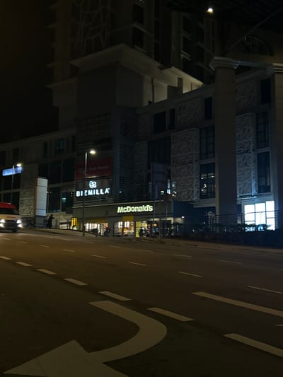 The image shows a street scene at night in Putrajaya, Malaysia. A multi-story building with a modern design is prominently featured on the left side of the frame. Signs for "Bizmilla" and "McDonald's" are illuminated on the building's facade. The road on the right side of the image is marked with white lines and a painted white arrow. A few people appear to be walking in front of the McDonald's. Several cars are on the street and driving in the road. The overall environment is dark, with the light coming from street lamps, building signs, and vehicle headlights.