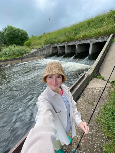 In the city of Bereznik, Russia, a woman is taking a selfie by a water canal. She is wearing a light-colored button-up shirt over a pink and gray top, a straw hat, and green boots. She is holding a fishing rod, and it appears she is fishing. The water in the canal is flowing rapidly, and there is a series of concrete culverts in the background. The sky is overcast, and there is a bird flying in the distance. The area around the canal is covered in grass and some concrete pathways.