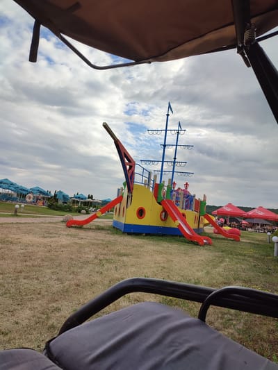 An outdoor shot in Shcherbani, Ukraine captures a colorful, ship-themed playground structure with red and yellow slides and blue masts, set on a dry, grassy field under a cloudy sky. In the background, rows of turquoise beach umbrellas and lounge chairs suggest a resort or recreation area. Several people are visible near the beach umbrellas, and a few are on the playground. The foreground shows the edge of a dark-colored seating area, possibly a swing or a shaded pavilion. Red Coca-Cola umbrellas are visible to the right, indicating a cafe or dining area.