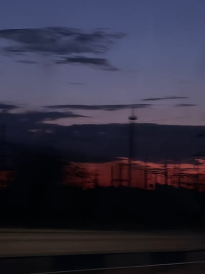 The image depicts a twilight sky with a gradient of deep blue to purple. Scattered, dark clouds are visible, some appearing wispy. In the mid-ground, silhouetted structures suggest industrial elements or infrastructure, possibly including power lines and a tall, slender tower. The lower part of the image is dominated by a dark, indistinct silhouette of trees or other vegetation, with a hint of red or orange light filtering through, suggesting a sunset or sunrise. A paved surface, possibly a road or highway, with faint white lines, occupies the very bottom of the frame, indicating a travel perspective. The overall impression is one of a rural or industrial outskirts scene at dusk. The location is identified as Shosseinyi, Russia.