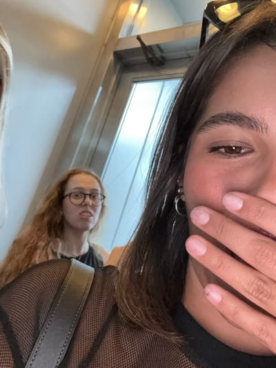 The image displays an indoor scene featuring three individuals. In the foreground, positioned to the right, is a subject with dark brown hair, whose right eye and eyebrow are visible. Their mouth is covered by their right hand, which exhibits pale, possibly chipped nail polish. They wear a black mesh top and silver hoop earrings, with sunglasses perched on top of their head. To the far left, a portion of another individual with blonde hair is visible. In the background, a woman with long, curly reddish-brown hair and eyeglasses is seen with an open mouth, revealing teeth, and an expression of surprise or distress. She is wearing a dark-colored top. The environment consists of light-colored walls and a glass door or window frame with a visible metal latch in the upper center-right. The specific location within Philadelphia, United States, cannot be identified from the visual data presented.