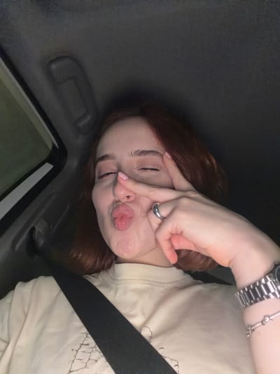 A young woman with red hair is in a car, wearing a seatbelt. She is making a peace sign with her hand over one eye and puckering her lips. She is wearing a silver watch and a silver ring. The image was taken at night. The location is Grigalati, Georgia.