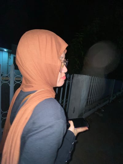 A young woman wearing a brown hijab and glasses is captured from the side, walking at night. She is holding a black smartphone in her right hand, seemingly engaged with it. Her attire consists of a dark-colored top, partially visible beneath the hijab and a portion of a brown scarf draped over her shoulder. The background reveals a dimly lit environment, suggesting an outdoor setting at night. A white fence runs along the right side of the image, and the ground appears to be a paved surface. There is a faint blur in the upper right quadrant of the image, possibly indicating a light source or atmospheric condition. The location is identified as Tanggul, Indonesia.
