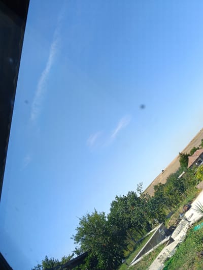The image is an angled shot of a bright blue sky with a few wispy clouds. In the lower right quadrant, a sloping, green landscape is visible, dotted with trees and a small building with a terracotta-tiled roof. A narrow, light-colored path runs through the grass, with some construction materials or debris near its edge. The scene appears to be outdoors, possibly in a rural or semi-rural setting outside of Suhache, Bulgaria.
