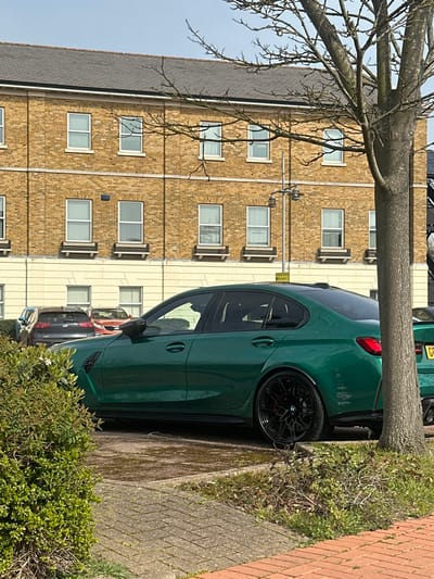 The scene depicts a parked dark green BMW M3 (G80 generation) sedan, positioned in a paved parking area on Pier Road, Gillingham, United Kingdom. The vehicle faces right and features black multi-spoke alloy wheels with red brake calipers, and tinted rear windows. Partially visible behind it are a dark grey SUV and an orange passenger vehicle. In the background stands a multi-story building constructed from light brown brick, featuring a dark grey roof and a cream-colored lower facade. The building has numerous white-framed rectangular windows, many fitted with vertical blinds. The windows on the second floor are accented by decorative planter boxes. To the right, a deciduous tree with bare branches partially obscures the building and a black metallic street light pole. A dense green bush occupies the foreground on the left. The ground consists of reddish block paving leading to the parking surface, which shows signs of dampness and moss. The sky is partially visible and clear, indicating daylight conditions consistent with late autumn, winter, or early spring. No persons are present.