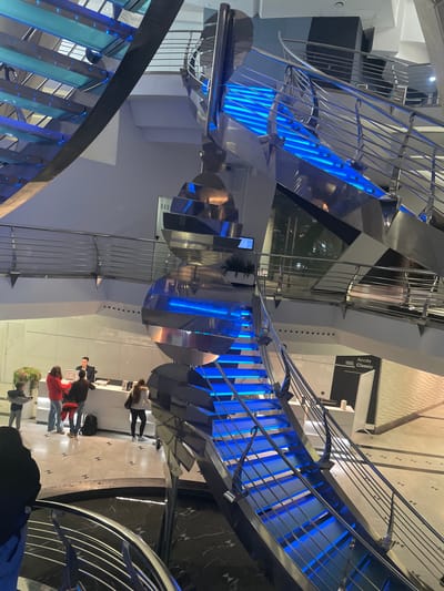The image depicts the interior of the Caldea thermal spa in Les Escaldes, Andorra. A modern, spiraling staircase with blue LED lighting dominates the scene. The staircase has metallic railings and is constructed with glass or acrylic risers. A sculptural element with rotating metallic disks hangs above the staircase.
Several people are visible on the lower level, including individuals standing in a queue near a reception desk. Signs with text are present on the wall. The surrounding environment includes a marble or patterned floor.