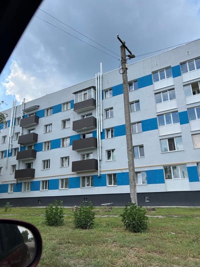 The image depicts a multi-story residential building in Staryi Saltiv, Ukraine, viewed from a slightly elevated position. The building facade is primarily light grey with prominent horizontal blue stripes at each floor level. Numerous windows with white frames are visible across all floors. Several open-air balconies with dark brown corrugated exteriors are present, along with one enclosed balcony on an upper floor. Multiple vertical silver-colored metallic pipes, consistent with exhaust or ventilation systems, are affixed to the building's exterior, running from lower levels to the roofline. A concrete utility pole stands in the mid-ground on the right, supporting overhead electrical lines and associated equipment. The foreground consists of a grassy area with three small, green leafy shrubs. The lower portion of the building's exterior is painted dark grey or black. In the bottom left corner, the red-painted side mirror of a vehicle is partially visible, reflecting a blurred background. The sky above is overcast with a mix of light and dark clouds. No people or active interactions are visible in the scene.