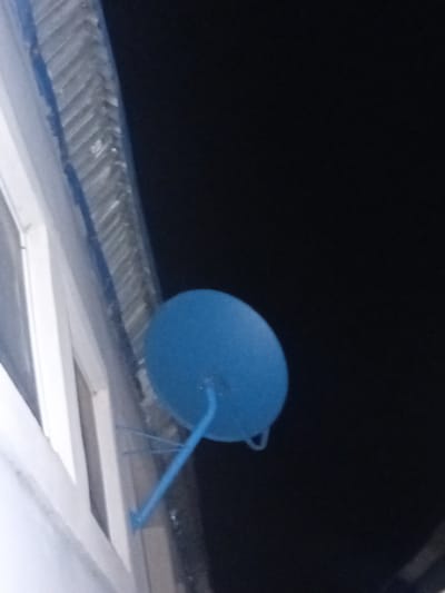 A blue satellite dish is mounted on the exterior wall of a building, with a dark sky in the background. The dish is circular and attached to a thin support arm. Part of the building's window frame, which is white, is visible on the left side of the image. The wall appears to have some textured siding or insulation. The image is taken at night, as indicated by the dark sky. The location is Pantinapu, Nigeria.