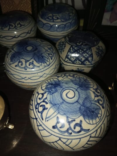 Five decorative porcelain balls with blue and white designs are arranged on a dark surface. Most are spherical, adorned with floral motifs and geometric patterns. One is shaped like a polygon. The designs feature varying blue shades, applied with a somewhat worn or vintage aesthetic. A partial view of an object with gold-colored detailing is visible on the left. A framed picture is partially visible in the background.