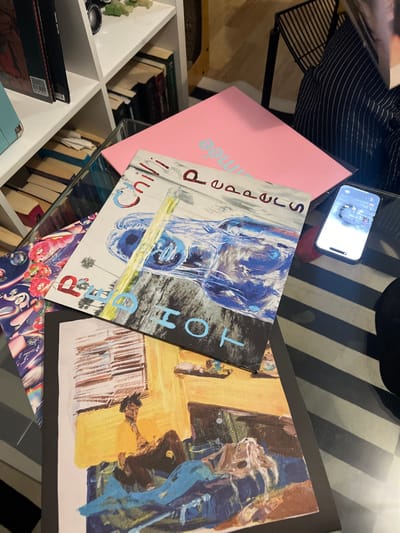A phone rests atop a stack including a pink book and Red Hot Chili Peppers’ “Dream Canteen” album. The stack sits on a glass-topped surface with books below. Another abstract painted canvas with yellow and blue hues rests to the side. The background includes a black and white striped surface and a white bookshelf filled with books.