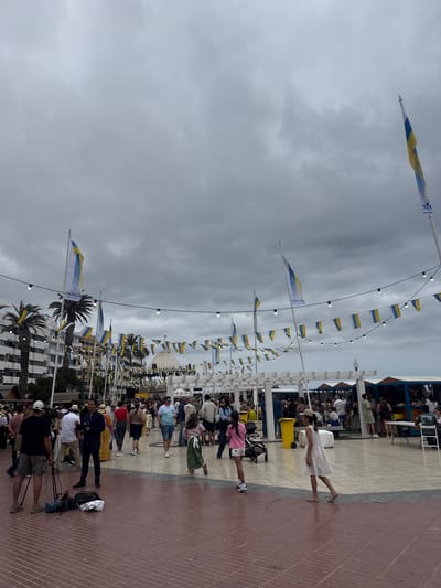The image shows a bustling public space in Arrecife, Spain, likely a promenade or plaza. The sky is overcast with grey clouds. Numerous people of varying ages and attire are present, some walking, others standing, and others seated at what appears to be an outdoor venue. A young woman wearing a white dress is walking across the pavement.

The area is decorated with rows of hanging flags bearing the colors blue and yellow, suspended between light poles with string lights. Behind the crowd are low-rise buildings and palm trees. The pavement is a mix of red-brown brick and lighter colored tiles, suggesting a well-maintained public area. A television camera and crew are also present.